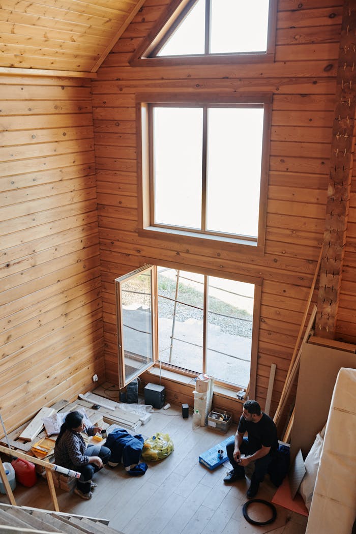 Interior of a rustic wooden cabin with two people resting amid construction materials, capturing a moment of pause.